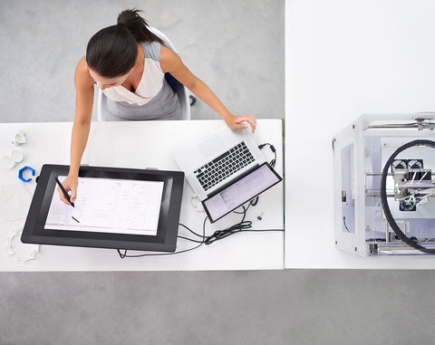 A woman at her desk, drawing on a large tablet connected to a laptop and 3D printer.